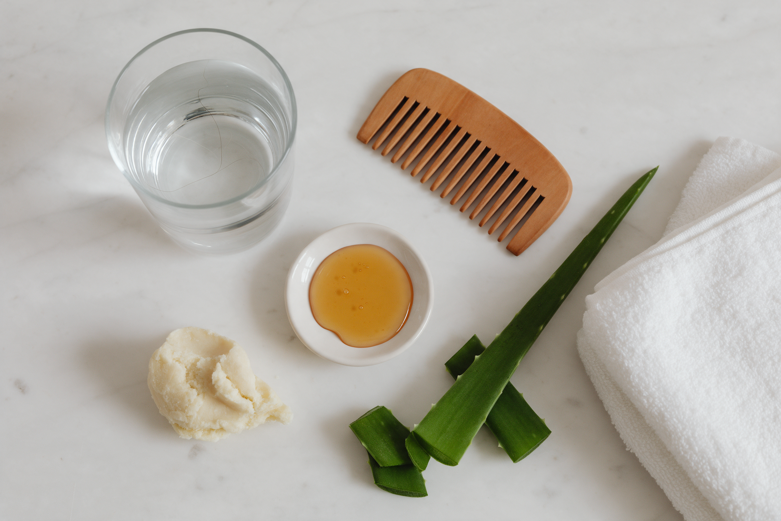 A glass of water, a wooden comb, honey, shea butter, aloe vera and a white towel on a marble counter